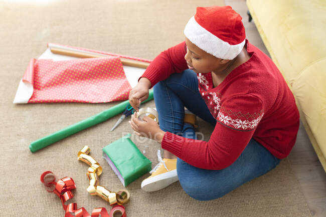 Feliz afro-americano plus size mulher em Papai Noel embrulhando presentes em casa. Natal, festa e conceito de tradição. — Fotografia de Stock