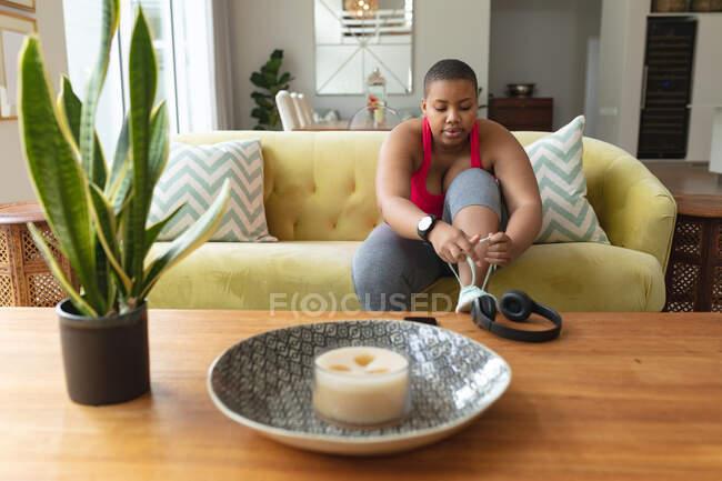 African american plus size woman in sports clothes sitting on sofa and tying shoes. fitness and healthy, active lifestyle. — Stock Photo