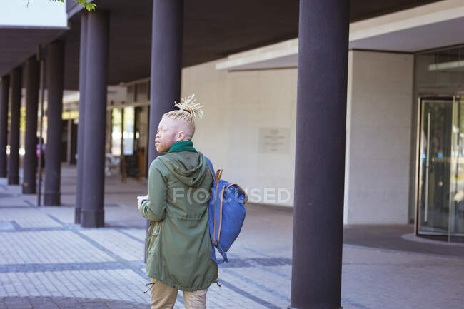 Pensativo albino hombre afroamericano con rastas caminando con un teléfono inteligente. nómada digital sobre la marcha, fuera y alrededor de la ciudad. - foto de stock