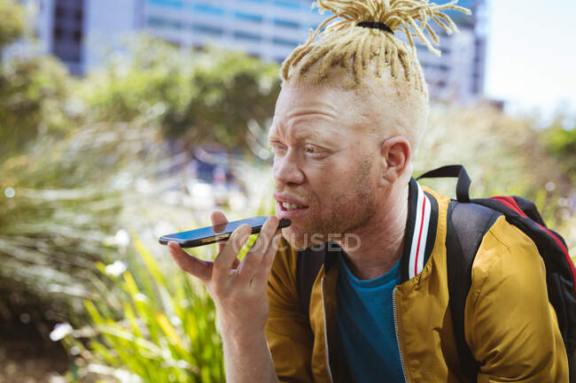 Heureux albinos homme afro-américain avec dreadlocks dans le parc parler sur smartphone. nomade numérique en déplacement, en déplacement dans la ville. — Photo de stock