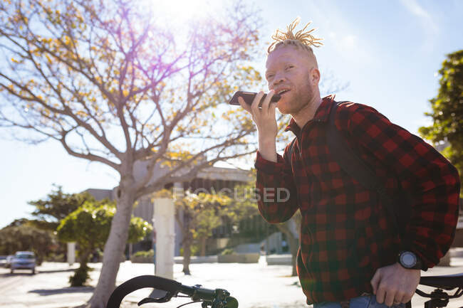 Feliz albino hombre afroamericano con rastas en el parque con la bicicleta hablando en el teléfono inteligente. nómada digital sobre la marcha, fuera y alrededor de la ciudad. - foto de stock