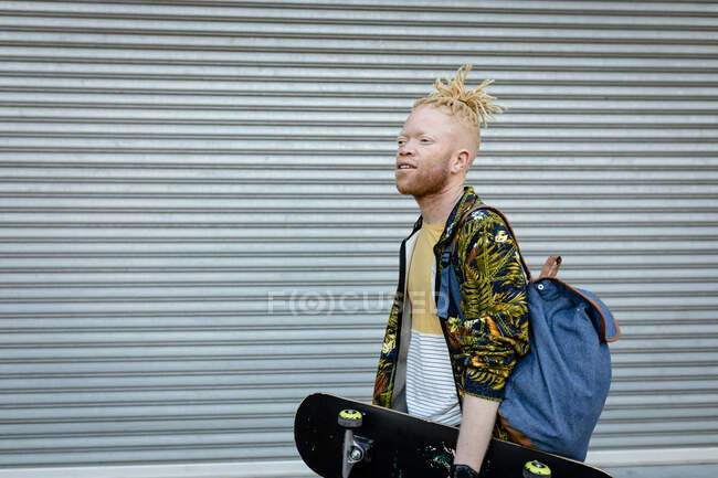 Heureux homme afro-américain albinos avec dreadlocks marchant tenant skateboard. on the go, out and about dans la ville. — Photo de stock
