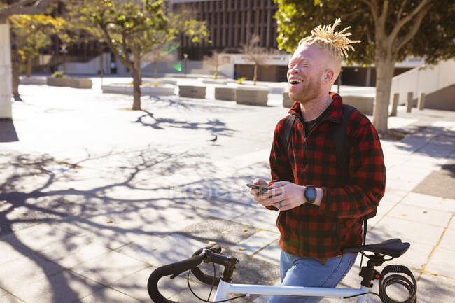 Homem americano africano albino feliz com dreadlocks em bicicleta usando smartphone. nômade digital em movimento, para fora e sobre na cidade. — Fotografia de Stock