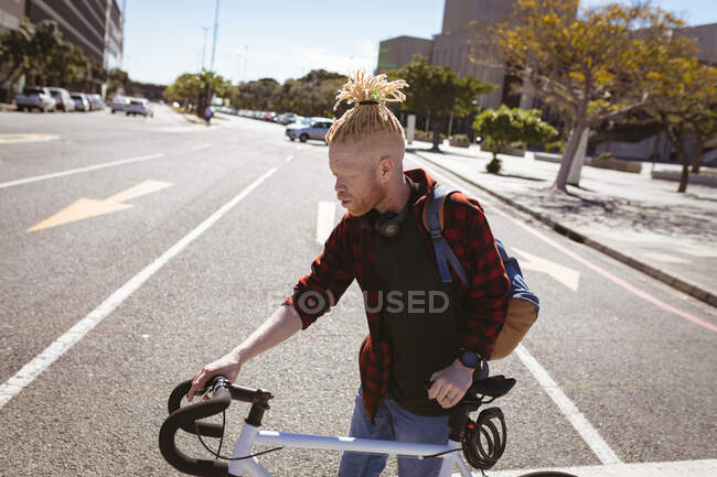 Thoughtful albino african american man with dreadlocks crossing road with bike. on the go, out and about in the city. — Stock Photo