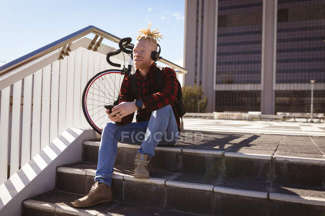 Homem americano albino atencioso afro-americano com dreadlocks usando fones de ouvido usando smartphone. nômade digital em movimento, para fora e sobre na cidade. — Fotografia de Stock