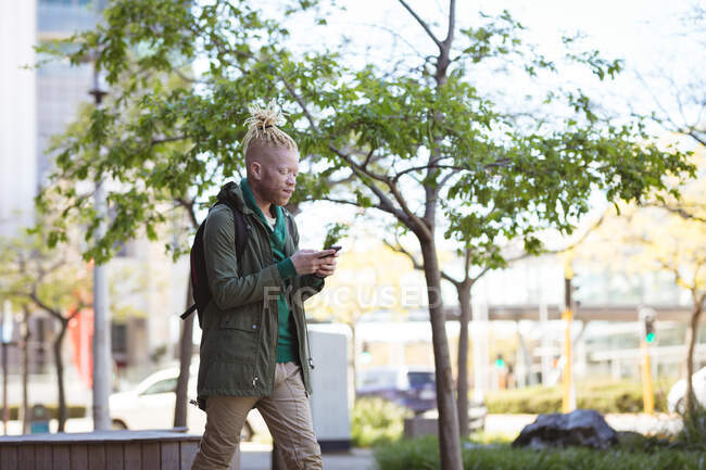 Pensativo albino hombre afroamericano con rastas caminando con un teléfono inteligente. nómada digital sobre la marcha, fuera y alrededor de la ciudad. - foto de stock
