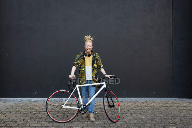 Retrato del sonriente hombre afroamericano albino con rastas en la calle con bicicleta. sobre la marcha, fuera y alrededor de la ciudad. - foto de stock
