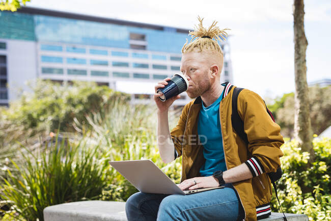 Nachdenklicher Albino-Afrikaner mit Dreadlocks, der im Park sitzt und Kaffee trinkt, mit Laptop. digitaler Nomade unterwegs, unterwegs in der Stadt. — Stockfoto