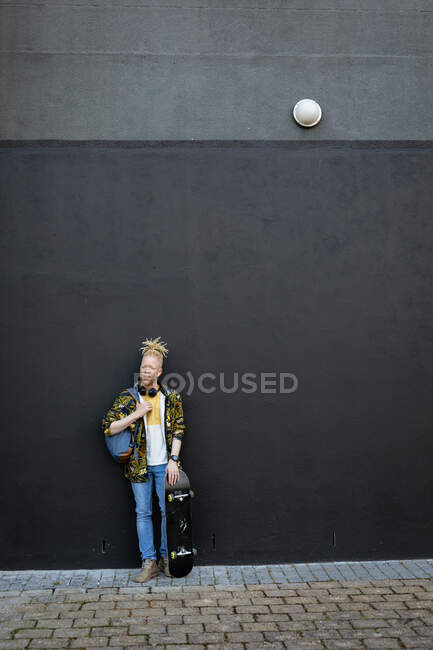 Retrato de albino feliz hombre afroamericano con rastas sosteniendo monopatín. sobre la marcha, fuera y alrededor de la ciudad. - foto de stock