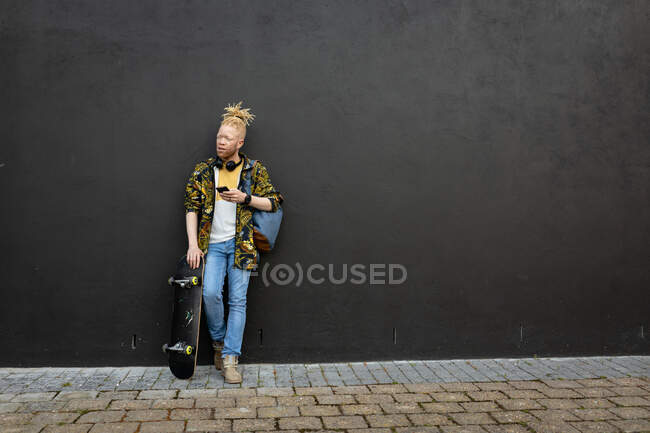 Nachdenklicher Albino-Afrikaner mit Dreadlocks, der mit dem Smartphone Skateboard hält. digitaler Nomade unterwegs, unterwegs in der Stadt. — Stockfoto