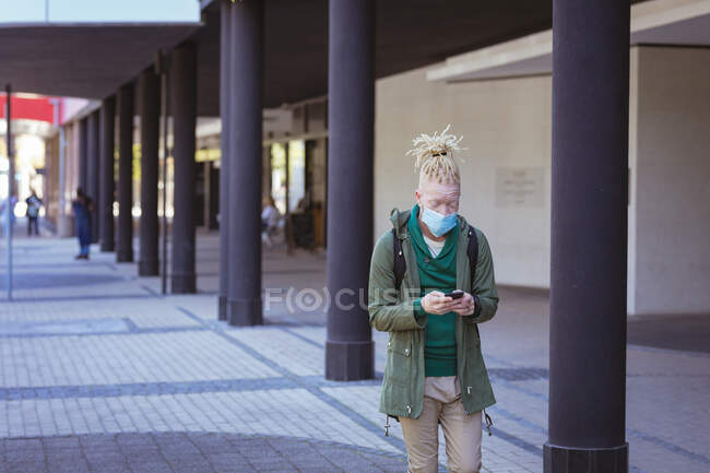 Albino homem americano africano com máscara facial e dreadlocks andando e usando smartphone. em movimento, para fora e sobre na cidade durante a pandemia covid 19. — Fotografia de Stock