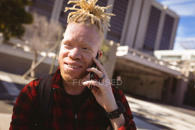 Glücklicher Albino-Afrikaner mit Dreadlocks im Smartphone-Gespräch. digitaler Nomade unterwegs, unterwegs in der Stadt. — Stockfoto