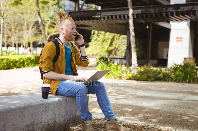 Pensativo albino hombre afroamericano sentado en el parque hablando en el teléfono inteligente utilizando el ordenador portátil. nómada digital sobre la marcha, fuera y alrededor de la ciudad. - foto de stock