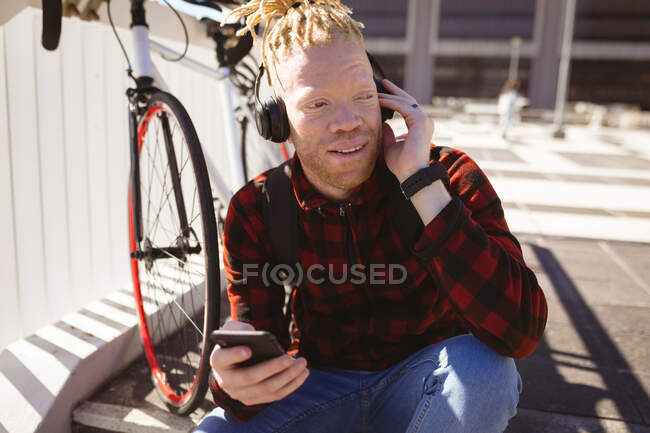Heureux homme albinos afro-américain avec dreadlocks portant un casque à l'aide d'un smartphone. nomade numérique en déplacement, en déplacement dans la ville. — Photo de stock