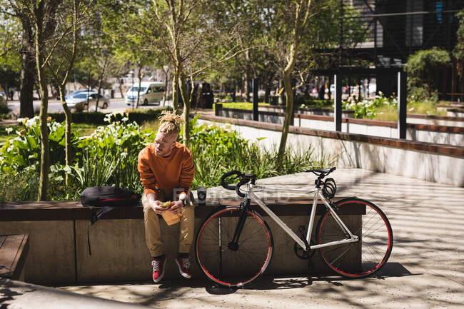 Albino-amerikanischer Mann mit Dreadlocks sitzt mit Fahrrad im Park und isst Sandwich. Unterwegs, unterwegs in der Stadt. — Stockfoto
