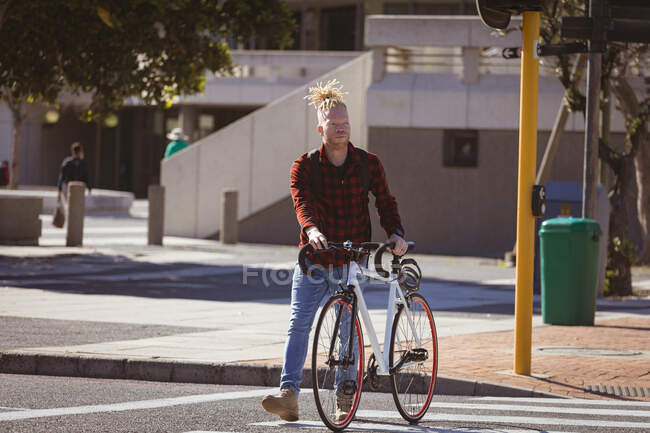 Продуманий альбіно-афроамериканський чоловік з дредлоками, що перетинають вулицю з велосипедом. на ходу, виходити і близько в місті . — стокове фото