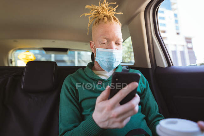 Albino african american man wearing face mask sitting in car using smartphone. on the go, out and about in the city during covid 19 pandemic. — Stock Photo