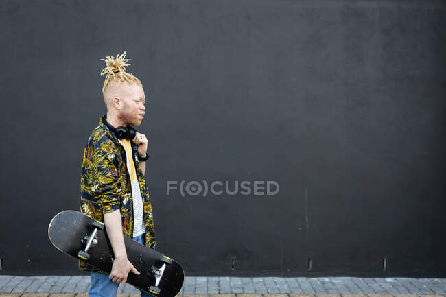 Feliz albino hombre afroamericano con rastas caminando sosteniendo monopatín. sobre la marcha, fuera y alrededor de la ciudad. - foto de stock