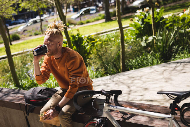 Albino uomo afroamericano con dreadlocks seduto nel parco con bici bere caffè. in movimento, in giro per la città. — Foto stock
