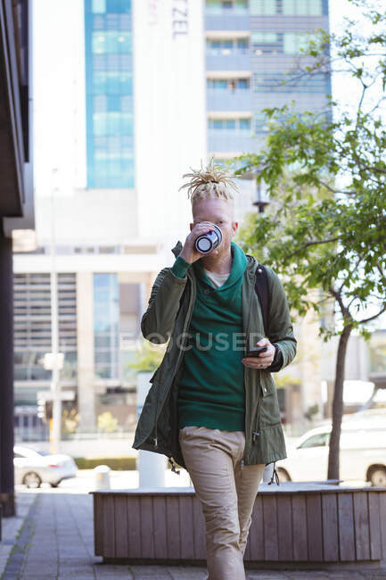 Thoughtful albino african american man with dreadlocks drinking coffee using smartphone. digital nomad on the go, out and about in the city. — Stock Photo