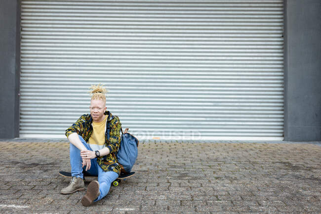 Retrato del reflexivo hombre afroamericano albino con rastas sentado monopatín. sobre la marcha, fuera y alrededor de la ciudad. - foto de stock