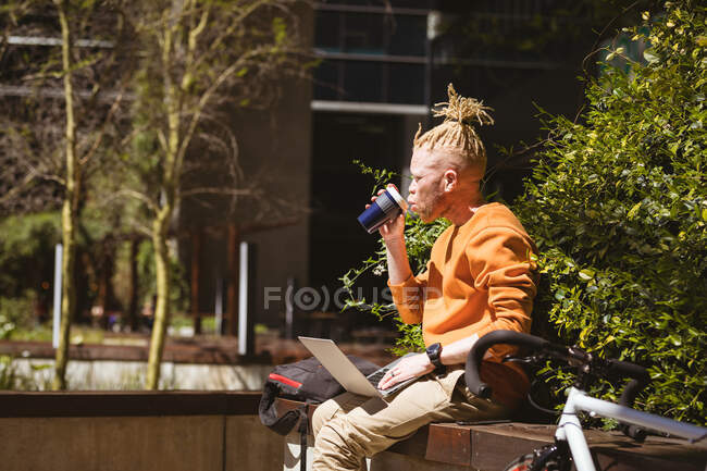 Thoughtful albino african american man with dreadlocks sitting in park drinking coffee using laptop. digital nomad on the go, out and about in the city. — Stock Photo
