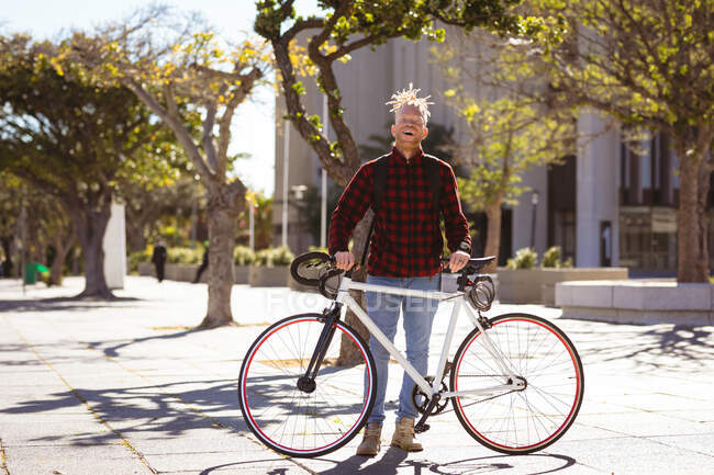 Retrato del sonriente hombre afroamericano albino con rastas en parque con bicicleta. sobre la marcha, fuera y alrededor de la ciudad. - foto de stock