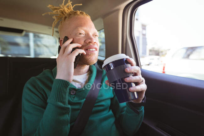 Feliz albino hombre afroamericano con rastas sentado en el coche hablando en el teléfono inteligente. nómada digital sobre la marcha, fuera y alrededor de la ciudad. - foto de stock