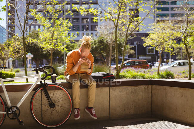 Albino african american man with dreadlocks sitting in park with bike eating sandwich. on the go, out and about in the city. — Stock Photo