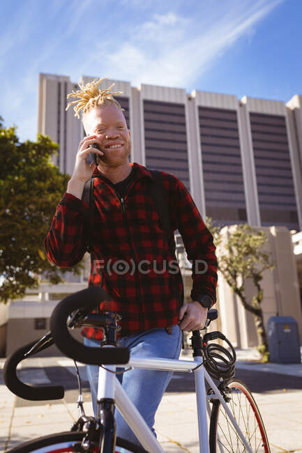 Retrato sonriente albino hombre afroamericano con rastas en el parque con bicicleta hablando en el teléfono inteligente. nómada digital sobre la marcha, fuera y alrededor de la ciudad. - foto de stock