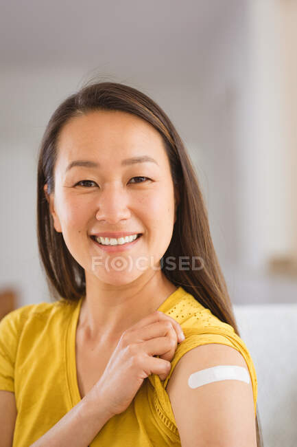 Portrait of happy asian woman sitting on sofa showing arm with plaster after vaccination. health and lifestyle during covid 19 pandemic. — Stock Photo