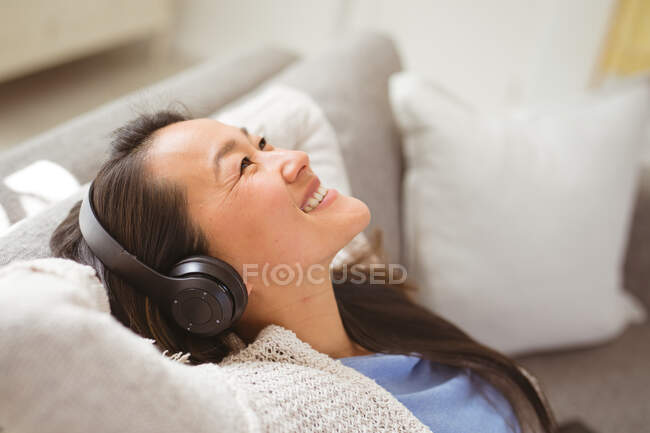 Feliz mulher asiática sentada no sofá com fones de ouvido em casa. estilo de vida e relaxar em casa com a tecnologia. — Fotografia de Stock