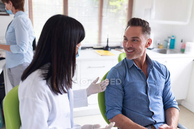 Dentiste naissante portant un masque facial et parlant avec un patient masculin à la clinique dentaire moderne. soins de santé et de la dentisterie. — Photo de stock