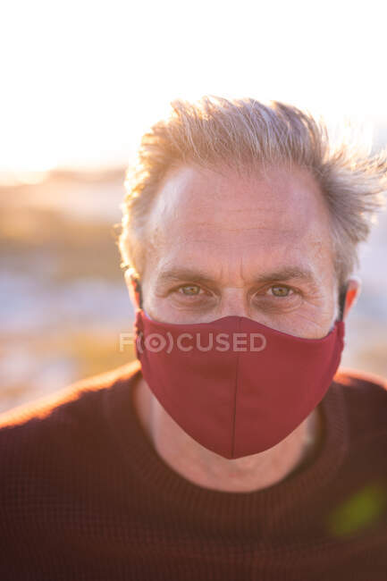 Portrait of caucasian man wearing face mask looking at camera at seaside. summer road trip and holiday in nature during covid 19 pandemic. — Stock Photo