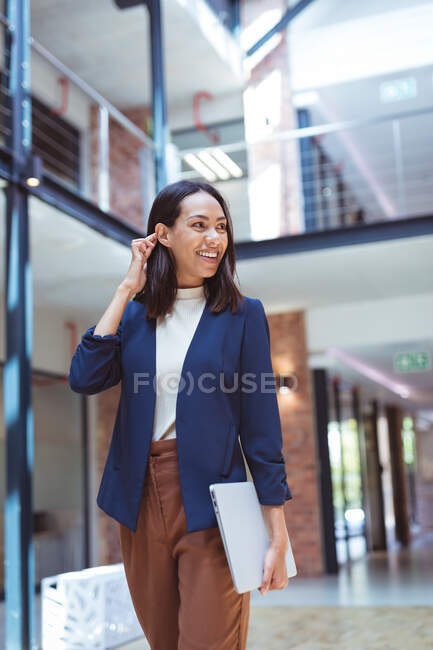 Biracial улыбается деловая женщина ходить с ноутбуком в современном офисе с коллегами в фоновом режиме. деловые и офисные рабочие места. — стоковое фото