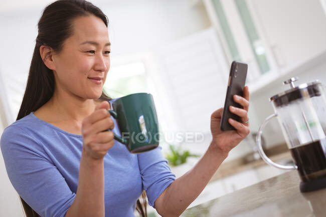 Щаслива азіатка п'є каву і використовує смартфон на кухні. спосіб життя і розслаблення вдома з технологіями . — стокове фото
