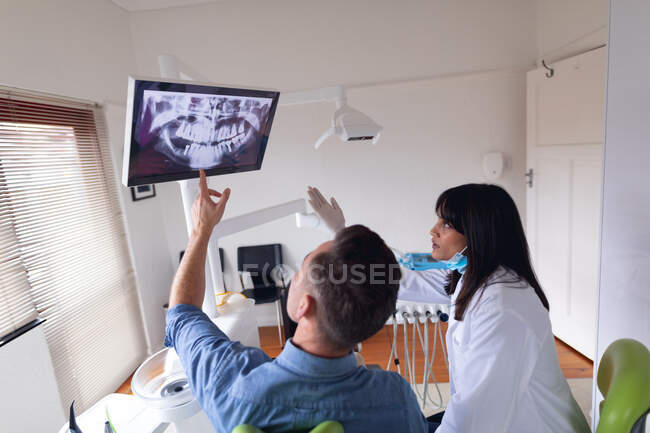 Dentista biracial usando máscara facial examinando dentes de paciente masculino na clínica odontológica moderna. serviços de saúde e odontologia. — Fotografia de Stock