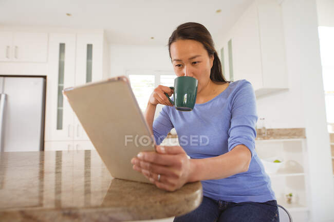 Фокус азиатской женщины пить кофе и использовать планшет на кухне. стиль жизни и отдых дома с технологией. — стоковое фото