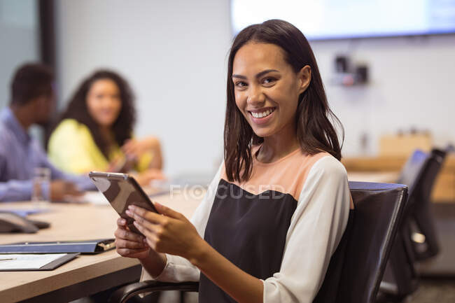 Retrato de mujer de negocios birracial sonriente usando tableta mirando a la cámara en la oficina moderna. negocios y oficina de trabajo. - foto de stock