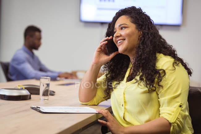 Biracial sonriente mujer de negocios hablando en el teléfono inteligente y sentado a la mesa en la oficina moderna. negocios y oficina de trabajo. - foto de stock