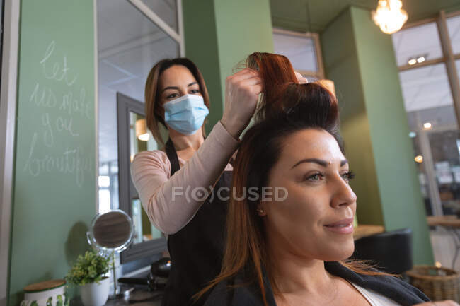 Peluquería femenina caucásica trabajando en peluquería usando mascarilla, poniendo rodillos de pelo en el cabello de cliente caucásica femenina. - foto de stock