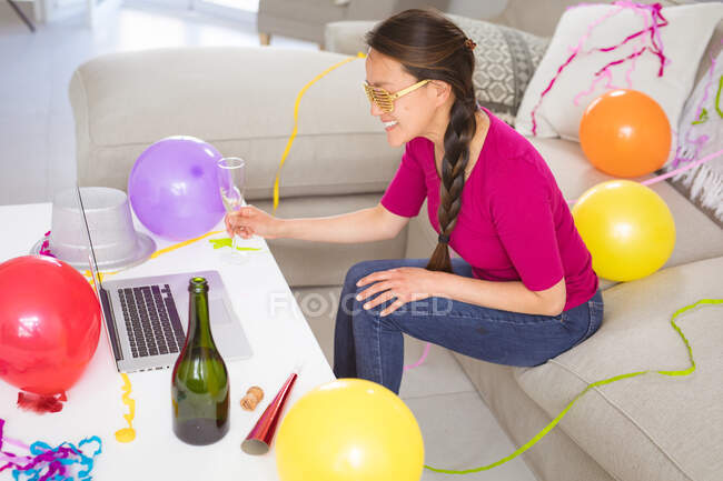 Счастливая азиатка поднимает бокалы с шампанским, делая видеозвонок на новогодний ноутбук. празднование дома с коммуникационными технологиями. — стоковое фото