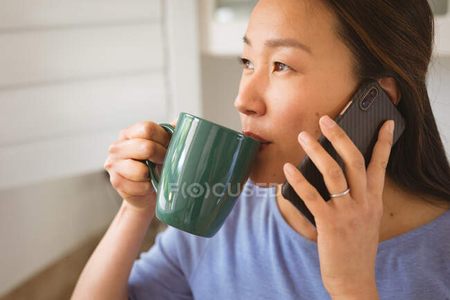 Retrato de mujer asiática feliz bebiendo café y usando smartphone en la cocina. estilo de vida y relajarse en casa con la tecnología. - foto de stock