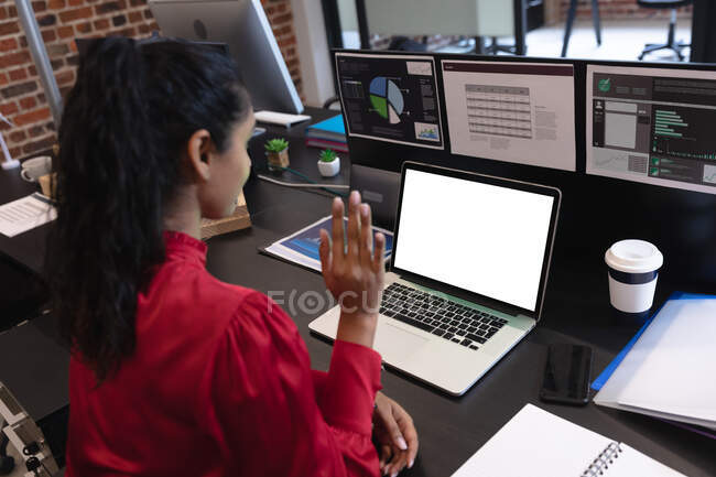 Gemischte Rassenfrau, die in einem lässigen Büro arbeitet, am Schreibtisch sitzt, einen Laptop benutzt und winkt. Soziale Distanzierung am Arbeitsplatz während der Coronavirus Covid 19 Pandemie. — Stockfoto