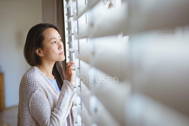 Perfil de mujer asiática reflexiva de pie en la ventana, mirando hacia el exterior. estilo de vida, ocio y pasar tiempo en casa. - foto de stock