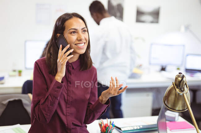 Feliz mujer de negocios birracial hablando en el teléfono inteligente con colegas en segundo plano en la oficina moderna. negocios y oficina de trabajo. - foto de stock