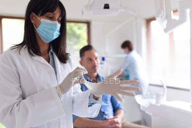 Dentista biracial feminina vestindo luvas médicas e paciente do sexo masculino esperando na clínica odontológica moderna. serviços de saúde e odontologia. — Fotografia de Stock