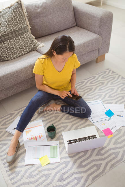Mujer asiática enfocada sentada en el suelo y trabajando remotamente desde casa con smartphone y laptop. oficina en casa y concepto de freelancing. - foto de stock