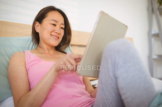 Heureuse femme asiatique allongée sur le lit, se reposant et utilisant une tablette. détente à la maison avec la technologie. — Photo de stock