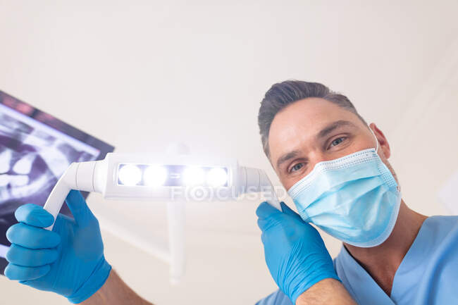 Dentista masculino caucasiano usando máscara facial segurando lâmpada na clínica odontológica moderna. serviços de saúde e odontologia. — Fotografia de Stock